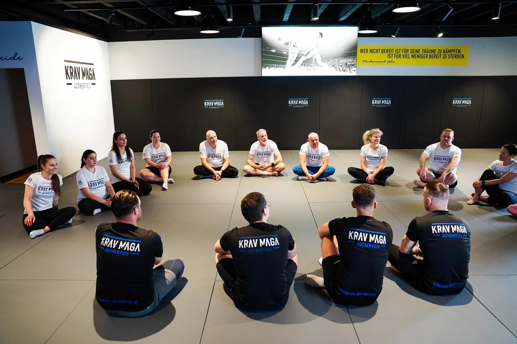 Krav-Maga-Kurs: Teilnehmer trainiert Tritttechnik am Pad, Trainer und Gruppe schauen zu